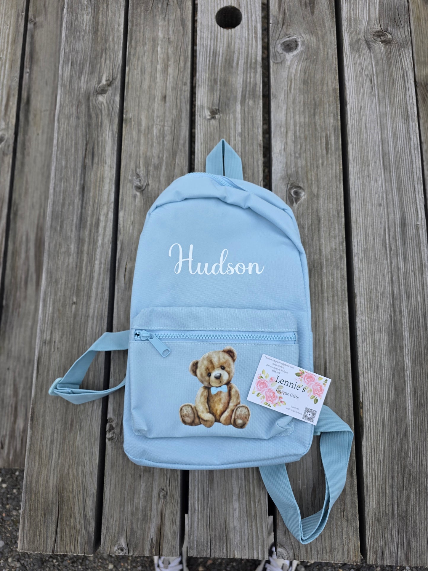 Light blue backpack with teddy bear design and 'Hudson' text on a wooden surface