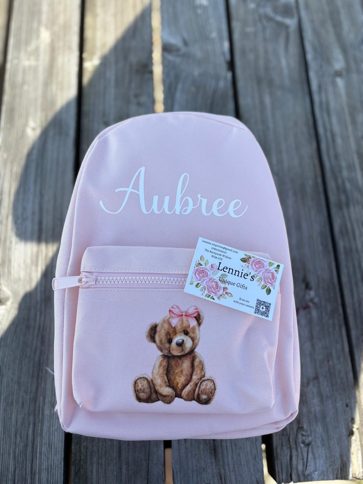Pink backpack with teddy bear design and 'Aubree' text on a wooden surface